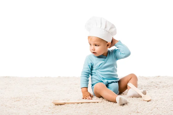 Niño pequeño en chefs sombrero sentado en la alfombra cerca de dos cucharas de madera grandes aislados en blanco - foto de stock