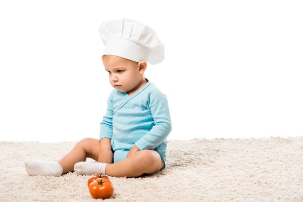 Serio bambino ragazzo in cuochi cappello seduto su tappeto vicino pomodori freschi isolati su bianco — Foto stock