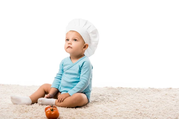 Carino bambino ragazzo in body blu e cuochi cappello seduto su tappeto vicino pomodori freschi isolati su bianco — Foto stock