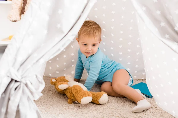 Cute toddler boy in blue bodysuit sitting on carpet in baby wigwam — Stock Photo