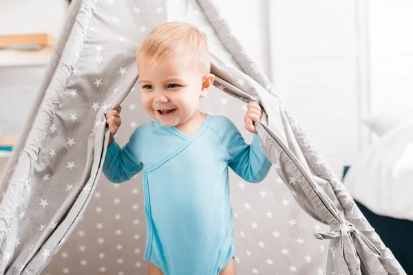 Close up view of smiling toddler boy in blue bodysuit standing in baby wigwam — Stock Photo