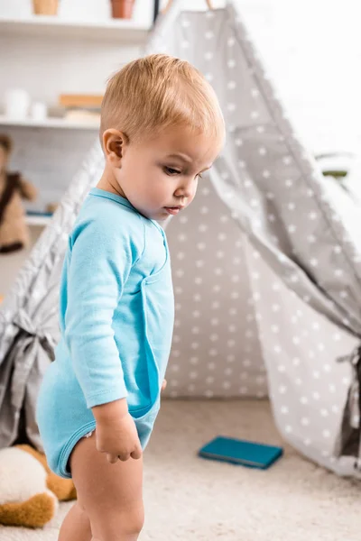 Close up view of cute toddler boy in blue bodysuit standing near grey baby wigwam — Stock Photo