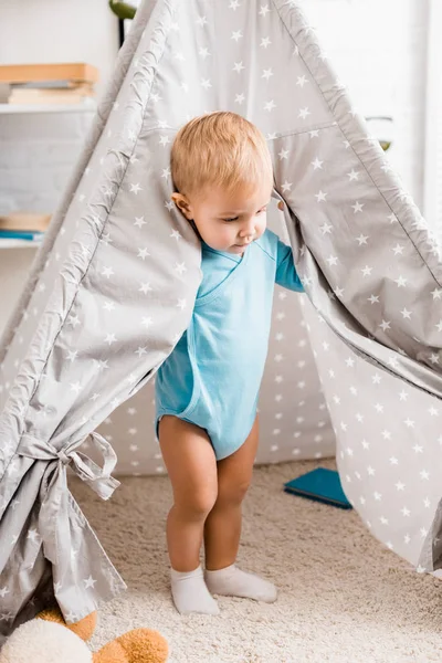 Cute toddler boy in blue bodysuit standing in grey baby wigwam on carpet — Stock Photo