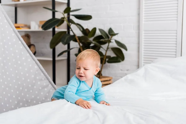 Sad baby in blue bodysuit standing near bed and crying in nursery room — Stock Photo