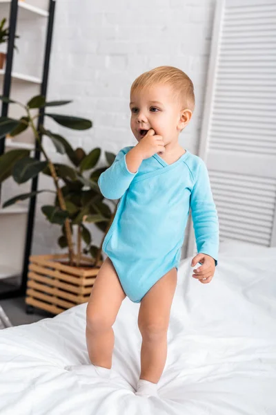 Cute baby putting finger in mouth and standing on bed in light room — Stock Photo
