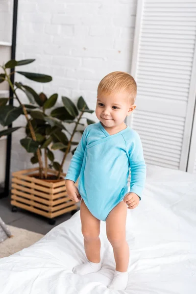 Bebé sonriente en traje de cuerpo azul de pie en la cama en la habitación del vivero - foto de stock