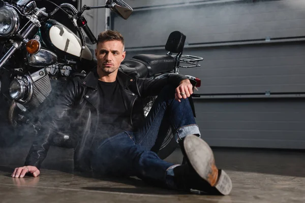 Handsome guy sitting on ground by motorcycle in garage — Stock Photo