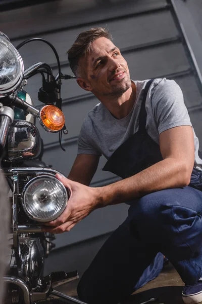 Smiling adult mechanic fixing motorcycle headlamp in garage — Stock Photo