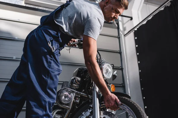 Smoking adult mechanic in overalls checking motorcycle tire in garage — Stock Photo