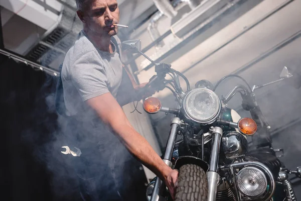 Cara fumar cigarro e verificar motocicleta roda dianteira na garagem — Fotografia de Stock