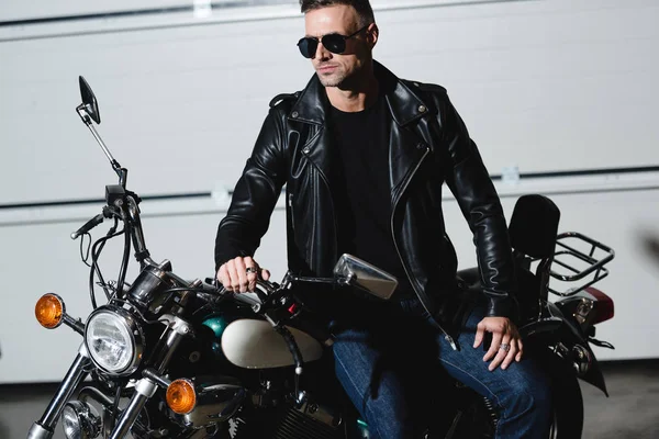 Bel homme classique en lunettes de soleil noires et veste en cuir debout à moto dans le garage — Photo de stock