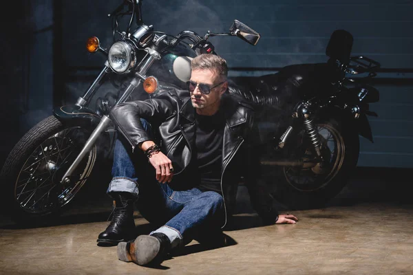 Handsome biker in sunglasses sitting on floor next to motorcycle in garage — Stock Photo