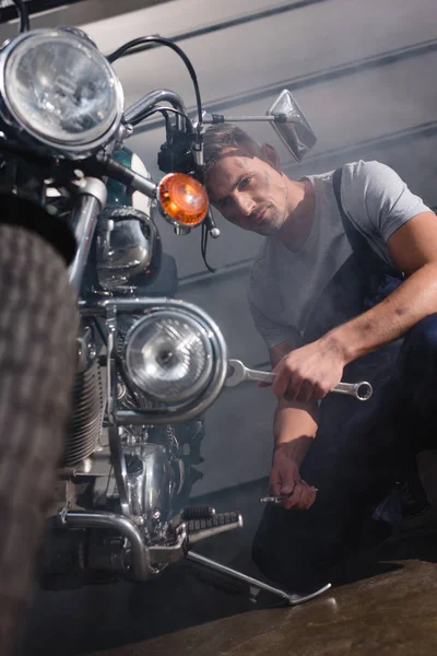 Schöner Mechaniker hält Schraubenschlüssel und fixiert Motorrad-Motor in Garage — Stockfoto