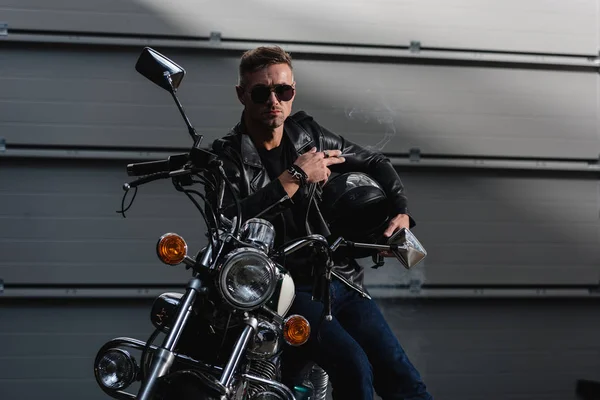 Chico guapo en gafas de sol negras y chaqueta de cuero sentado en motocicleta en el garaje - foto de stock