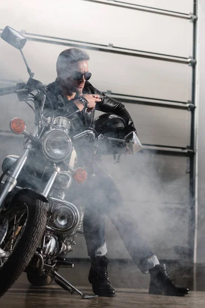 Guapo jinete en gafas de sol negras apoyado en la motocicleta en el garaje - foto de stock