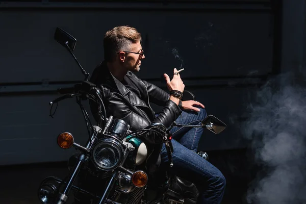 Handsome biker in sunglasses sitting on motorbike in garage — Stock Photo