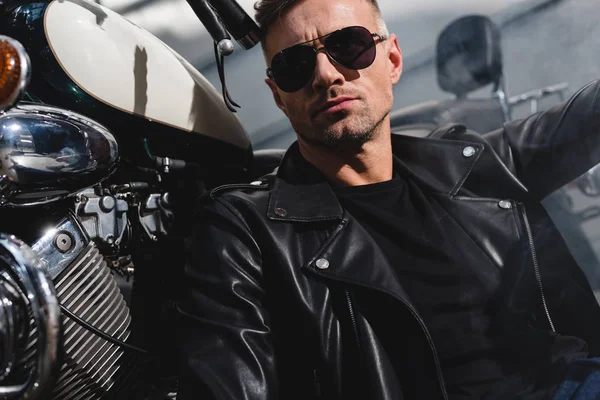 Handsome classic guy in sunglasses sitting by motorcycle in garage — Stock Photo