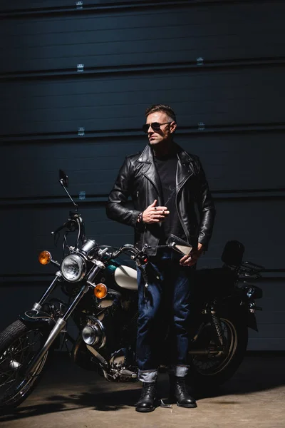 Chico clásico en gafas de sol de pie junto a la motocicleta en el garaje y fumar cigarrillos - foto de stock