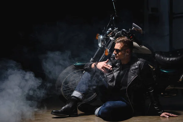 Beau motard en lunettes de soleil noires assis sur le sol à côté de la moto dans le garage — Photo de stock