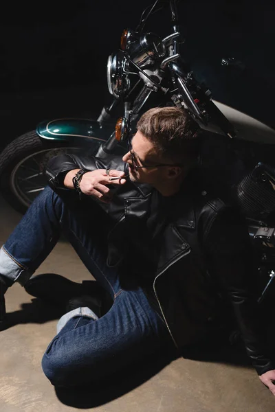 Handsome man in black sunglasses and leather jacket sitting on floor next to motorcycle — Stock Photo
