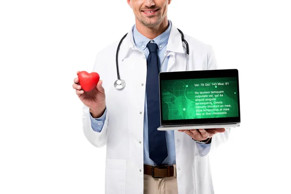 Vista recortada del médico sonriente sosteniendo el modelo de corazón y el ordenador portátil con datos de salud en la pantalla aislada en blanco, concepto de salud del corazón - foto de stock
