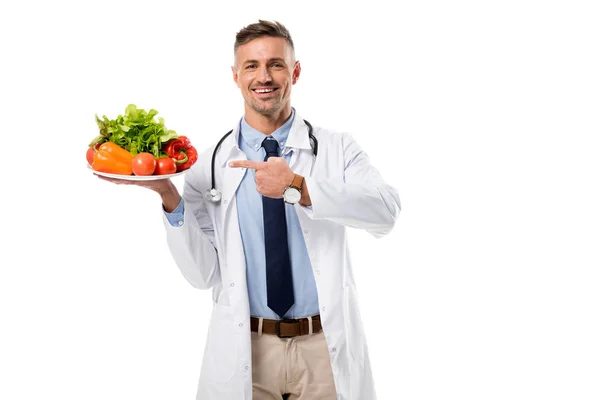 Lächelnder Arzt zeigt mit dem Finger auf Teller mit frischem Gemüse und Grün, isoliert auf weißem, gesundem Ernährungskonzept — Stockfoto