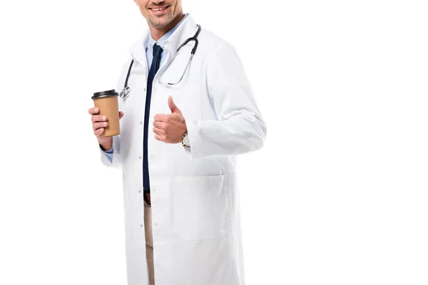 Обрізаний вид усміхненого лікаря, який тримає каву, щоб піти і зробити великий палець знак вгору ізольовано на білому — стокове фото