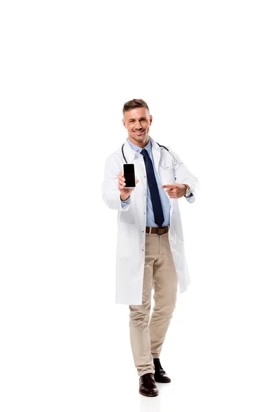 Médico sonriente apuntando con el dedo al teléfono inteligente con pantalla en blanco aislado en blanco - foto de stock