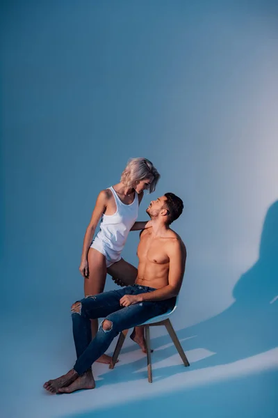 Joven mujer apasionadamente mirando al hombre sentado en la silla con sombras sobre fondo azul oscuro - foto de stock