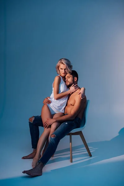 Attraktive Frau und Mann sitzen auf Stuhl, umarmen sich und schauen vor dunkelblauem Hintergrund weg — Stockfoto
