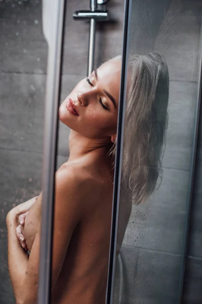 Красива молода жінка, що покриває груди і позує, приймаючи душ у ванній — стокове фото