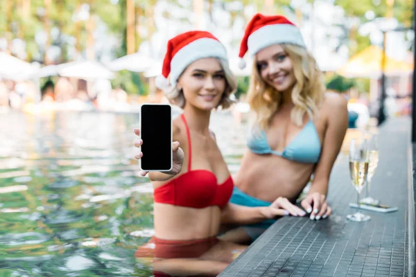 Smiling women in swimming suits and santa claus hats showing smartphone with blank screen while relaxing in swimming pool — Stock Photo