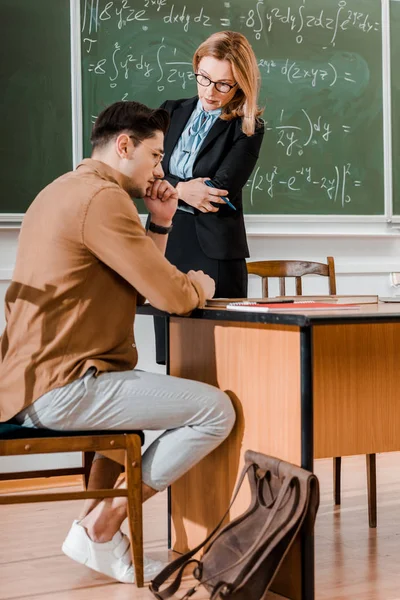 Professeure debout avec les bras croisés et regardant un étudiant réfléchi en classe — Photo de stock