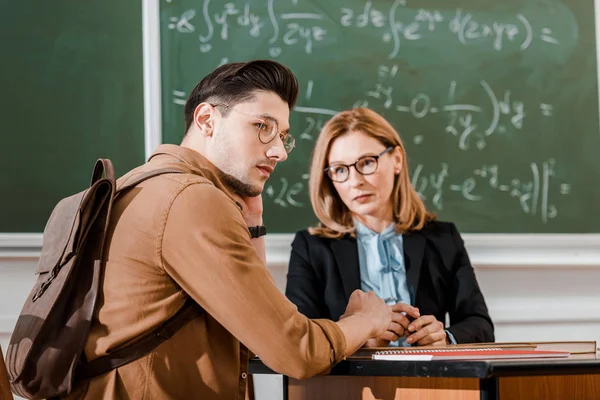 Female teacher looking at young student in classroom with chalkboard on background — Stock Photo