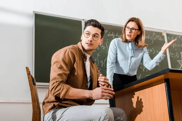 Dissatisfied female teacher standing near chalkboard and speaking with student in classroom — Stock Photo