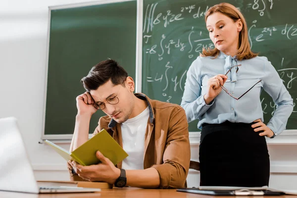 Estudiante en gafas sentado en el escritorio y mirando el cuaderno cerca del profesor - foto de stock