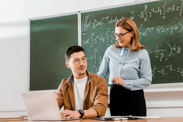 Jovem estudante usando laptop e sentado perto do professor em sala de aula — Fotografia de Stock