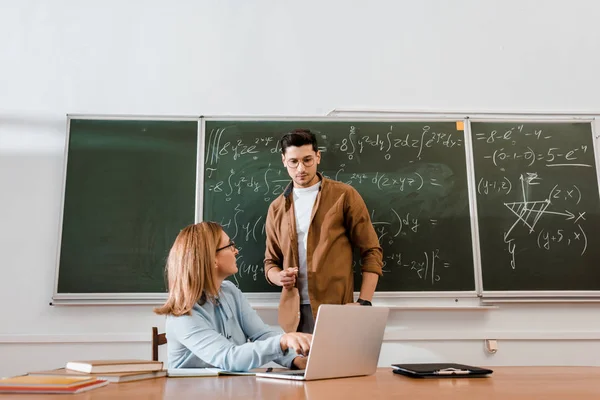 Female teacher looking at student and pointing at laptop in class — Stock Photo