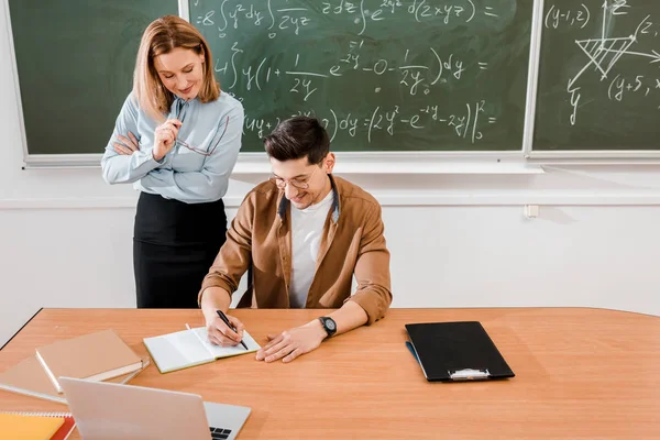 Estudiante en gafas wtiting en cuaderno cerca del profesor en el aula - foto de stock