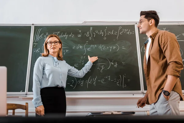 Enseignant féminin debout dans des lunettes et montrant des équations près de l'étudiant — Photo de stock