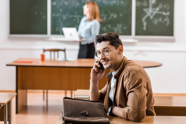 Estudiante hablando en el teléfono inteligente en el aula con el profesor en segundo plano - foto de stock