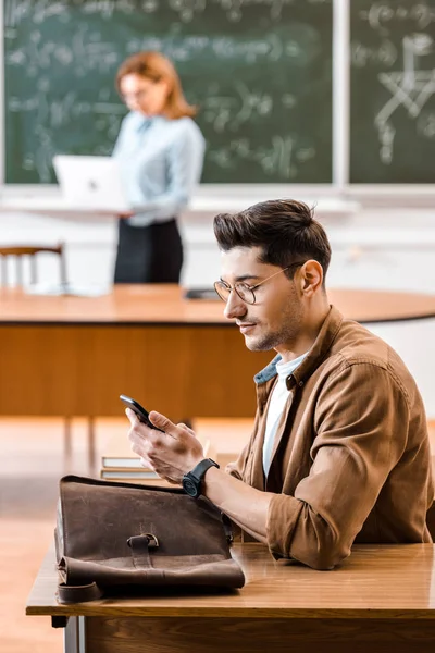 Focus selettivo dello studente utilizzando lo smartphone in classe con insegnante sullo sfondo — Foto stock