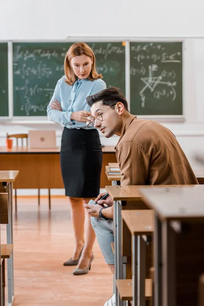 Dissatisfied female teacher with arms crossed looking at male student cheating during exam in classroom — Stock Photo