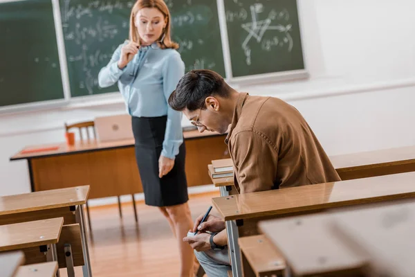 Selective focus of dissatisfied female teacher looking at male student cheating during exam in classroom — Stock Photo