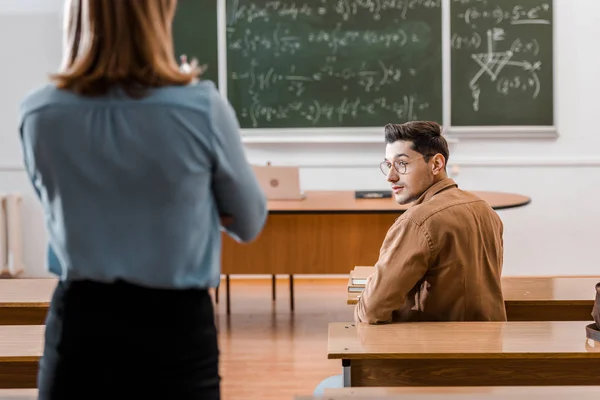 Vista posterior de la profesora mirando a un estudiante sentado en el escritorio durante la lección en el aula - foto de stock