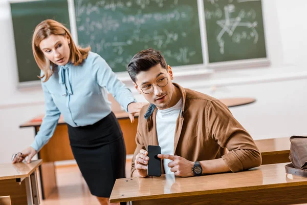 Insegnante di sesso femminile guardando studente maschio in possesso di smartphone durante la lezione in aula — Foto stock