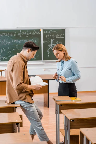 Estudiante masculina sosteniendo libro cerca de profesora en el aula - foto de stock