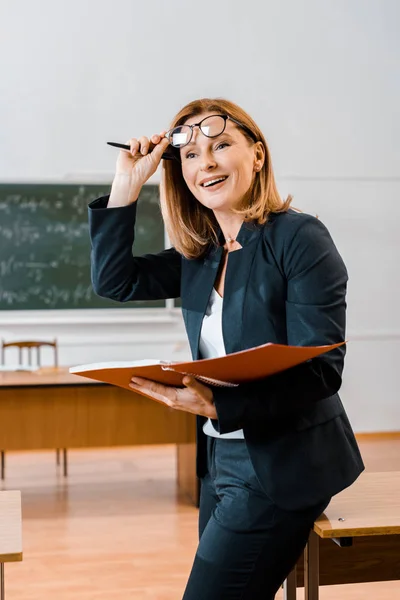 Beautiful smiling female teacher in formal wear and glasses holding notebook in classroom — Stock Photo