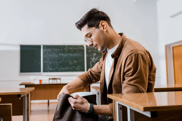 Focado estudante masculino em óculos sentado na mesa e segurando saco de couro em sala de aula — Fotografia de Stock