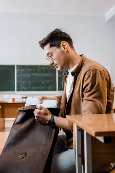 Концентрований студент чоловічої статі в окулярах, сидячи за столом і упаковуючи блокноти в класі — стокове фото